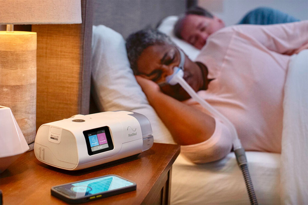 A couple sleeping in bed, with the woman in the foreground wearing a ResMed CPAP mask connected to a machine on the nightstand.