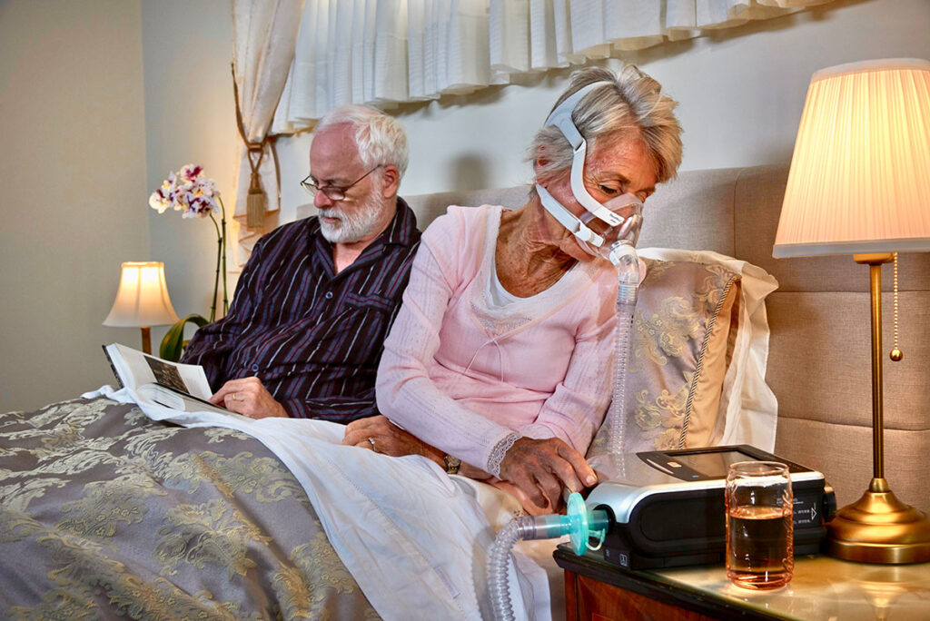 An older couple in bed at home, woman is using a ventilator and the man is reading a book.