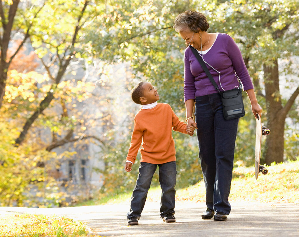 A woman with a portable oxygen concentrator shoulder bag carries a skateboard as she walks on a path holding hands with a young boy.