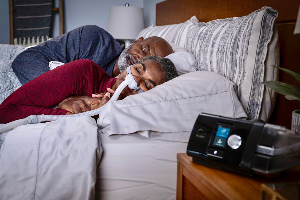 A man and woman are sleeping in bed at home, with the woman in the foreground wearing a ResMed BiPAP mask connected to a machine on the nightstand.