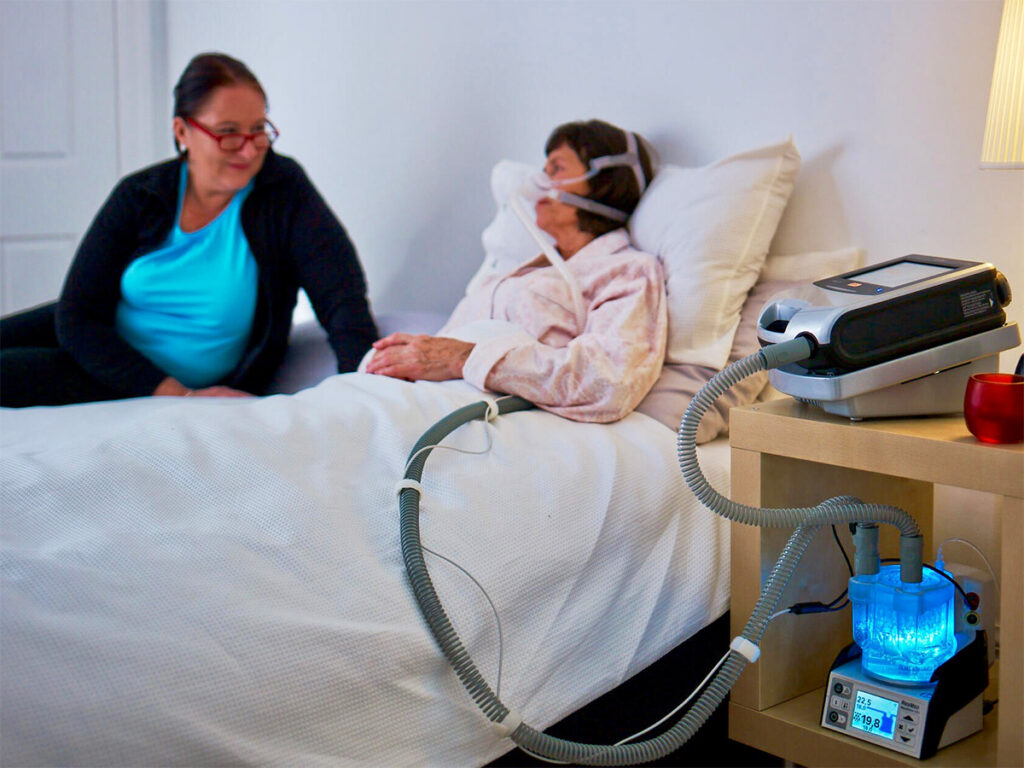 A woman lies in bed at home, hooked up to a ventilator on a nightstand, while a caregiver sits beside her.