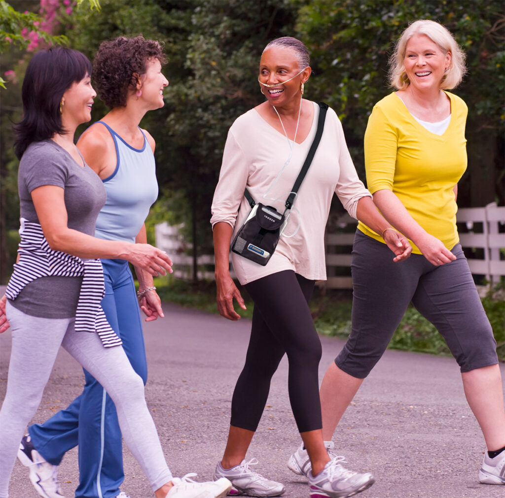 Three women walking down a path, smiling, wearing athletic clothing; woman in center wears portable oxygen concentrator with shoulder bag.