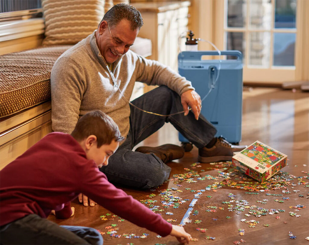 A man and a boy are sitting on the floor, building a puzzle. The man is using a stationary oxygen concentrator.