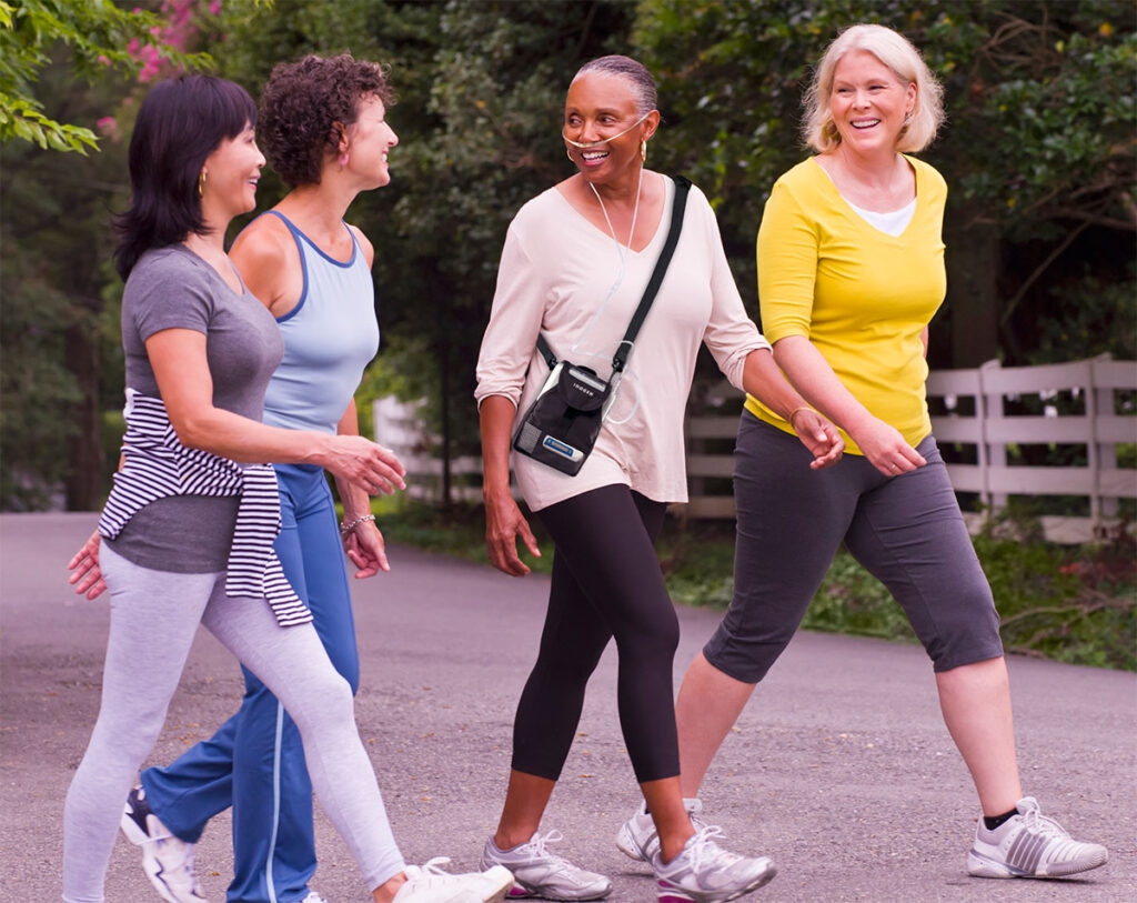 Three women walking down a path, smiling, wearing athletic clothing; woman in center wears portable oxygen concentrator with shoulder bag.