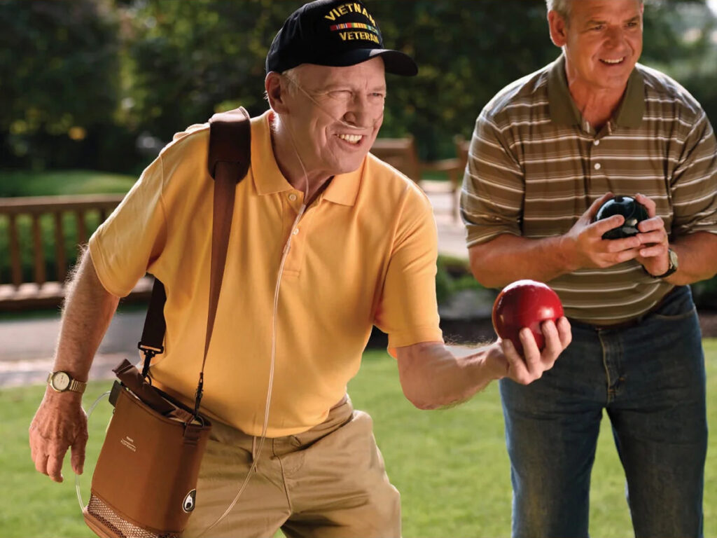 Two men are smiling together at a park holding balls; one man wears a portable oxygen concentrator with a shoulder bag.