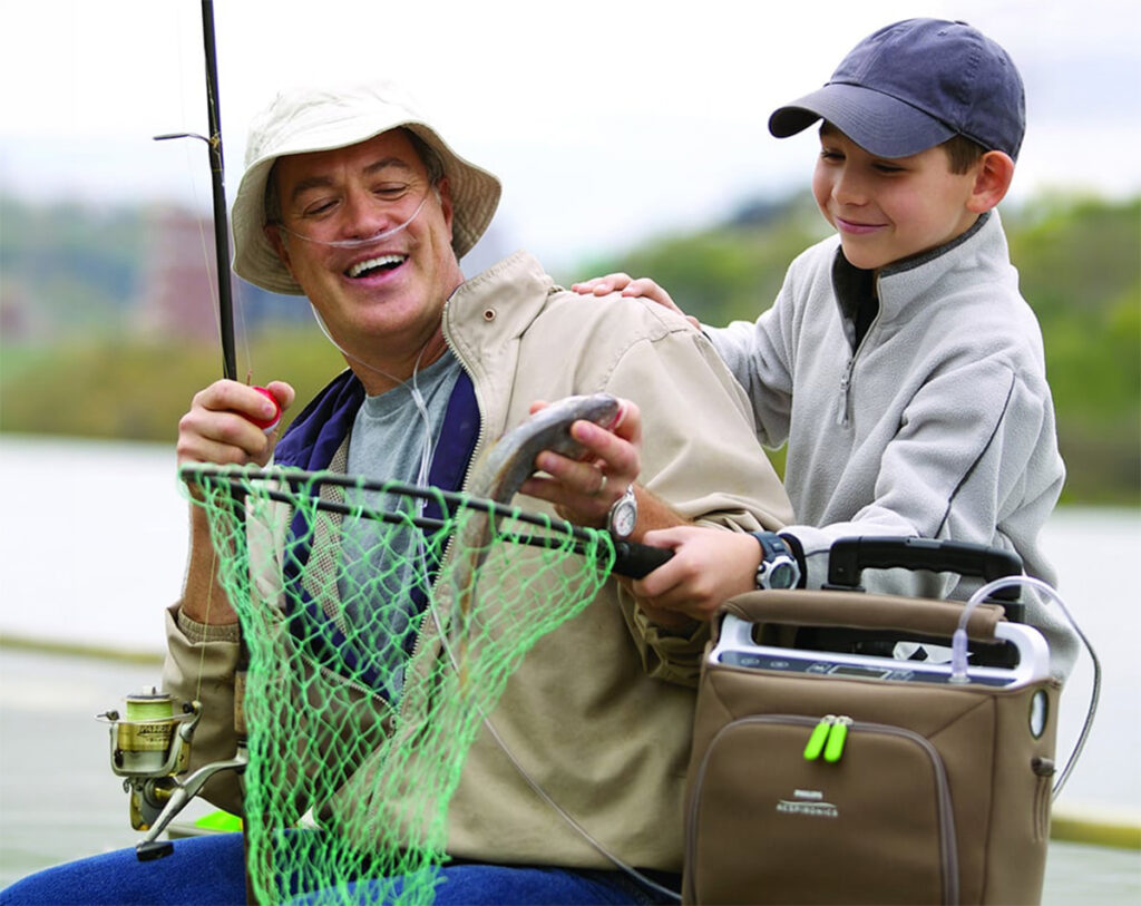 A man and a boy are sitting in a fishing boat, smiling at each other, holding a fish; the man uses a portable oxygen concentrator.