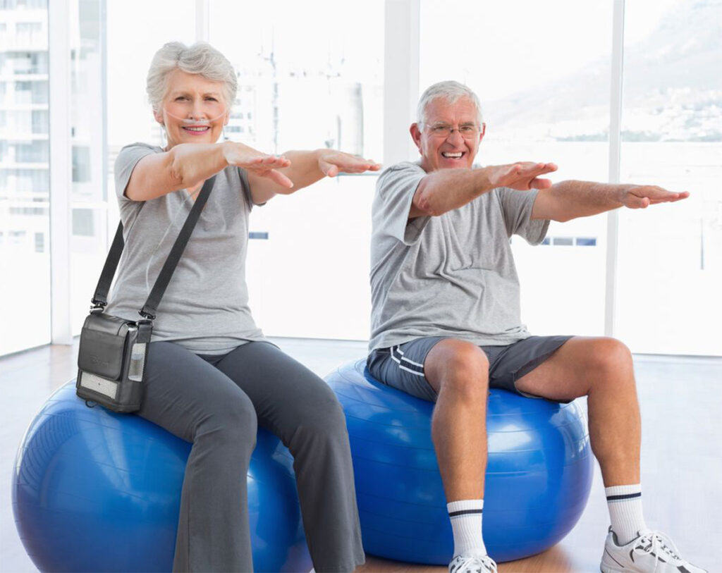An older man and woman are smiling, sitting on exercise balls in a gym. The woman is wearing a portable oxygen concentrator with a shoulder bag.