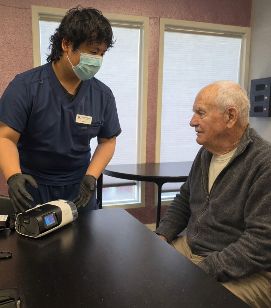SleepCare Source technician wearing gloves and a mask is with an older man at a table, setting up a CPAP machine.